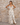 Matte All Dresses collection image – model wearing a cream tropical-print maxi dress in front of a textured beige stone wall in warm natural sunlight