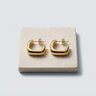 Chunky square gold hoop earrings with a polished mirror finish displayed on a natural stone tray against a soft gray background.