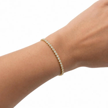 Gold bezel tennis bracelet with round clear stones worn on wrist, delicate minimalist jewelry styling, polished gold finish, and clean white studio background.
