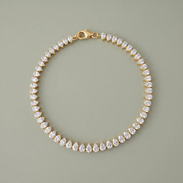 Gold teardrop tennis bracelet with sparkling clear stones and a polished silver clasp on a soft sage gray background, photographed in a clean luxury jewelry editorial style with refined reflections and minimal shadows.
