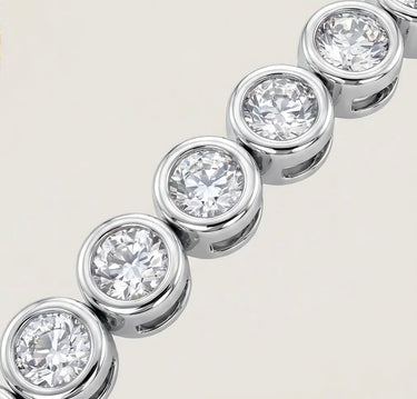 Close-up detail of a silver bezel tennis bracelet with round clear stones, polished silver settings, and reflective luxury finish on a warm light gray background.
