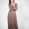Model wearing Caracol Dress in mocha brown, featuring long sleeves, V-neckline, and ruffled hem.
