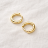 Cassis Huggie Earrings – small polished gold huggie hoop earrings with a smooth minimalist design, photographed on a soft neutral background.
