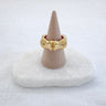 A gold-colored ring displayed on a beige mount, set against a white background.