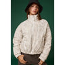 Woman wearing light neutral quilted puff jacket with high neck and elastic cuffs against dark green background.