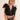 Woman wearing the Cyrene Top, a black feather-trimmed sequin crop top with bold texture and deep V neckline, photographed on a warm neutral backdrop.