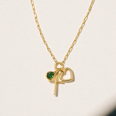 Debon Gold Charm Necklace close-up on neutral background showing gold cross, open heart, and emerald green stone details.