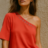 Woman wearing a coral one-shoulder top with embellishments against a beige wall.