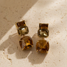 Encia Stud Earrings gold earrings with double-drop warm crystal stones on neutral textured background, showing faceted stones and soft golden tones.