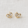 Kuga Stud Earrings featuring gold heart-shaped settings with clear crystals on a soft beige fabric background.