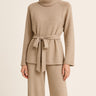 Woman wearing Hailee Lounge Sweater Set in beige, featuring knit turtleneck top with waist tie and wide-leg pants.
