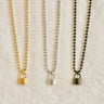 Lock Me Necklaces in gold, silver, and black with padlock pendants displayed on a beige fabric background.