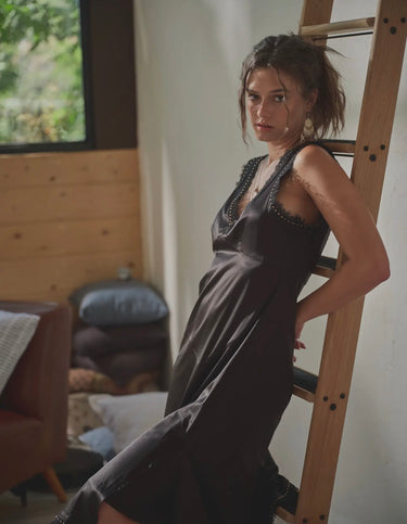 Woman wearing black satin asymmetric dress with lace neckline and delicate studs, seated by wooden ladder.
