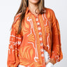 Maréa Blouse sunset-orange statement blouse with artistic tropical-inspired print, button front detail, and relaxed fit long sleeves, styled on model with white pants against clean studio background.