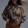 Flowy animal-print blouse with golden accents in brown and black tones, shown on model styled with hat and brown pants — Marisma Blouse by Matte.
