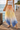 Merlot Pants, flowy resort pants with peach to yellow to ocean blue gradient worn on model walking on wooden dock near water showing lightweight fabric and wide-leg movement