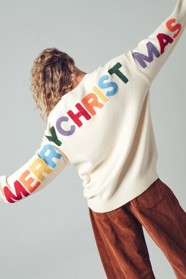 Model wearing ivory knit sweater with colorful MERRY CHRISTMAS lettering on sleeves and back.
