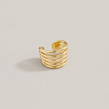 Mima Ear Cuff in polished gold featuring a wide layered open-band design with sleek stacked lines, styled as a bold modern statement ear cuff on a soft neutral background.