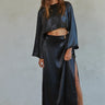 A woman wearing a long-sleeve crop satin top in black, paired with a high-waisted skirt with a side slit. 
