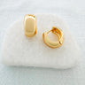 A pair of Natalia gold-plated huggie earrings displayed on a white cushion.