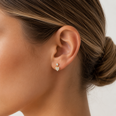 Woman wearing delicate gold pavé heart stud earrings with a minimalist fine jewelry design, styled on the ear in a close-up beauty portrait with soft neutral lighting.
