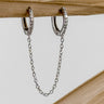 A pair of gold-plated huggie earrings with a chain connecting them, displayed against a wooden background.