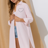 Woman wearing a light pink oversized shirt with a heart design, standing against a beige curtain.