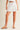 Ragit Short white tailored pleated shorts front view, featuring high-rise waist, clean structured pleats, and relaxed summer silhouette styled with neutral sandals.