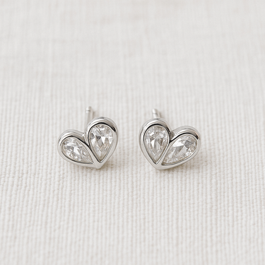 Kuga Stud Earrings featuring silver heart-shaped settings with clear crystals on a soft beige fabric background.