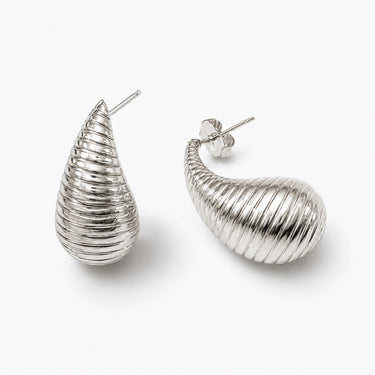 Silver ribbed teardrop statement earrings with sculptural curved silhouette and high-shine polished finish on a light neutral background.
