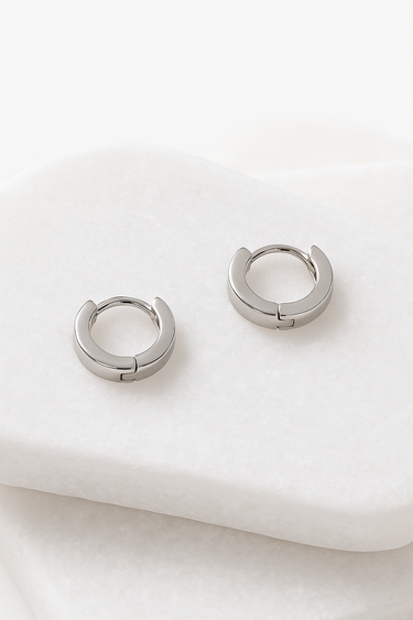 Silver minimalist huggie hoop earrings laying flat on white marble background – Stella Huggie Earrings by Matte