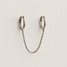 Wagu Huggie Earrings in silver with a connecting chain displayed on a light beige fabric background.