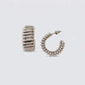 Polished silver ribbed hoop earrings with a chunky sculptural silhouette and high-shine finish on a soft light gray background.
