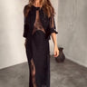 A woman wearing a black maxi dress with sheer details and a beaded design, standing in a room with a minimalist decor.