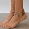 Silver beaded anklets on a person's feet with a neutral background