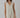 A sleeveless, beige linen dress with fringed detailing at the V-neckline and a raw hem, worn by a model.