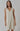 A sleeveless, beige linen dress with fringed detailing at the V-neckline and a raw hem, worn by a model.