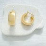 Ixelles Huggie Earrings in polished gold finish resting on a white stone against neutral fabric background.