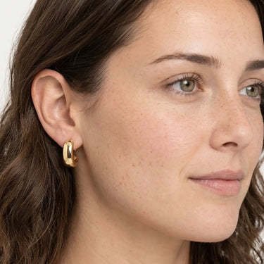 Model wearing Kayseri Hoop Earrings featuring polished gold-plated hoop earrings with a smooth domed rectangular front, rounded open hoop silhouette, lightweight design, high-shine finish, and a modern everyday look.