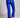 Model wearing cobalt blue slim pants with coated finish and white top — modern city look, back view.