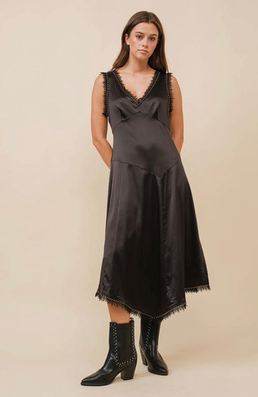 A woman wearing a black satin sleeveless dress with a V-neckline, lace trim, and stud embellishments. The dress has a flowy silhouette and an asymmetrical hem.