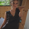 A woman standing next to a ladder, wearing a black satin sleeveless dress with a V-neckline and lace trim, paired with black boots.