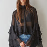 Woman wearing the Maruk Blouse in black, featuring a lightweight sheer fabric, relaxed silhouette, elegant high neckline, dramatic tiered bell sleeves, and flowing design styled with blue denim jeans.
