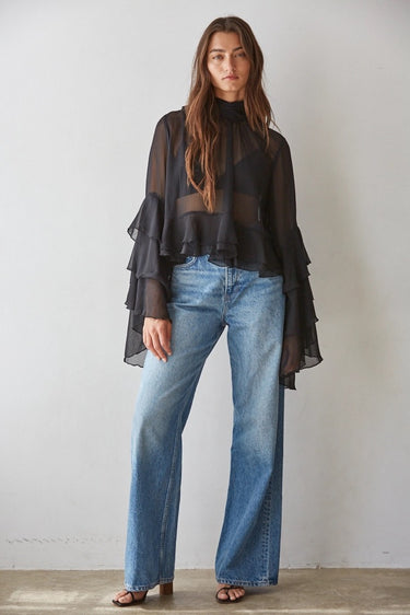 Full-body view of a model wearing the black Maruk Blouse with sheer fabric, cascading layered bell sleeves, relaxed fit, and light-wash denim for an elevated everyday outfit.