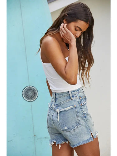 Model wearing the Minuett Top in white styled with distressed light-wash denim shorts, highlighting the asymmetrical neckline, ribbed stretch fabric, and effortless summer styling.