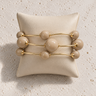 Minutti Bracelet in neutral beige featuring polished round beads and slim gold-tone bars in a sculptural multi-row design displayed on a cream jewelry cushion.