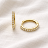 Naia Huggie Earrings – slim gold huggie earrings with pearl detailing, photographed on a soft neutral fabric background.