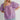 Model wearing the Nimes Top — lilac short-sleeve blouse with square neckline, puff sleeves, and textured stitching, styled with denim shorts for a soft bohemian look.