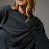 Woman wearing the Roux Top in charcoal black with sculpted ruching and dramatic draped detail, styled in soft natural light on a neutral background.