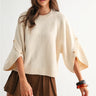 Sienu Sweater in cream featuring a relaxed oversized knit silhouette with cape sleeves, decorative button details, and a soft draped fit paired with a brown mini skirt.
